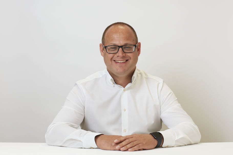 A person sits smiling at a white table, wearing glasses and a white shirt, with arms resting on the surface against a plain white background.