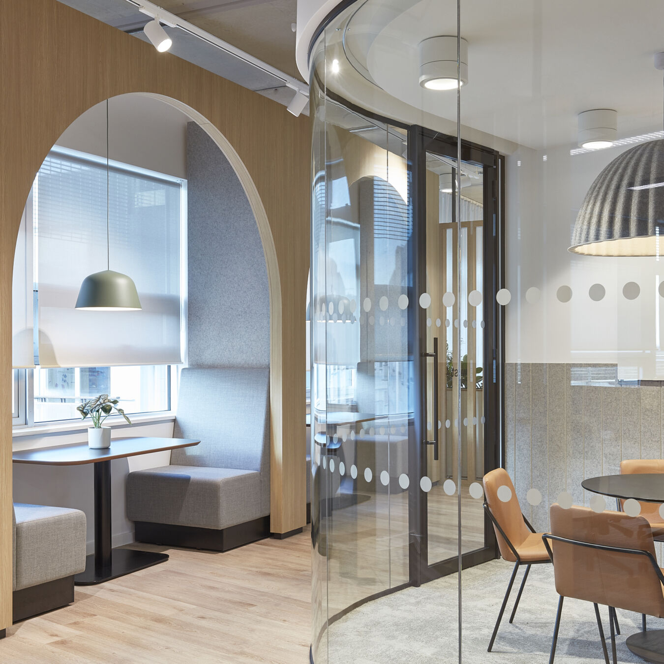 Modern office space with curved glass walls, featuring a round table with orange chairs. Booth seating is under an arched alcove with a pendant light