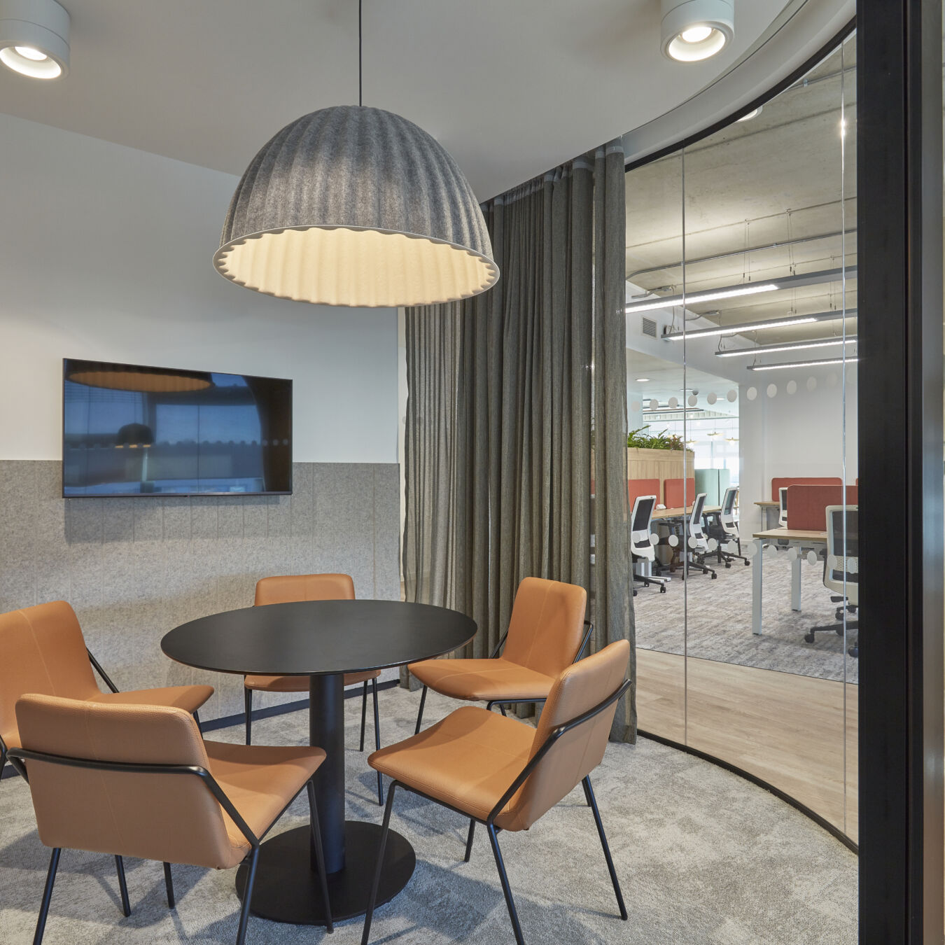 Small meeting room with round table, four tan chairs, large pendant light, and a wall-mounted screen, adjacent to an open office space