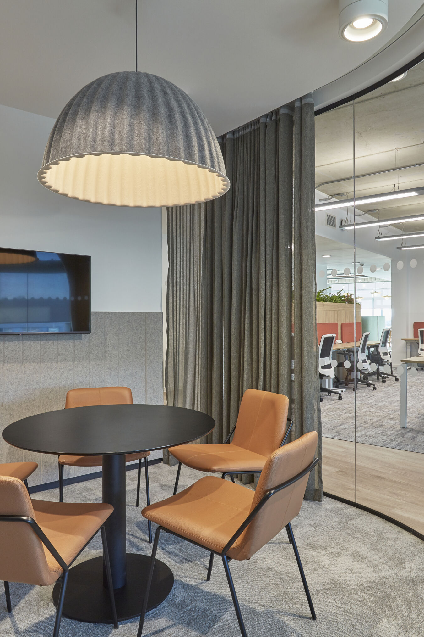 Small meeting room with round table, four tan chairs, large pendant light, and a wall-mounted screen, adjacent to an open office space