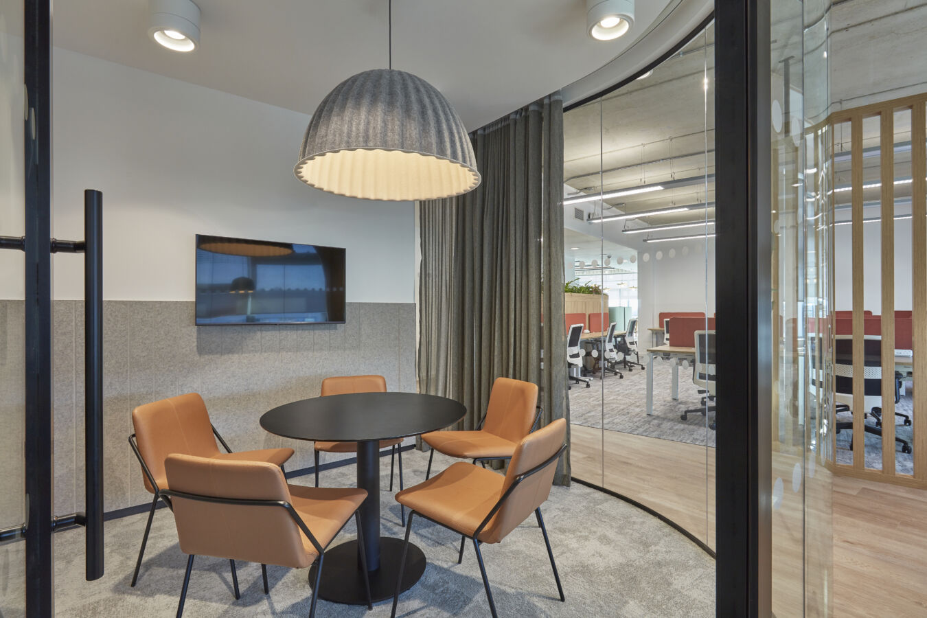 Small meeting room with round table, four tan chairs, large pendant light, and a wall-mounted screen, adjacent to an open office space