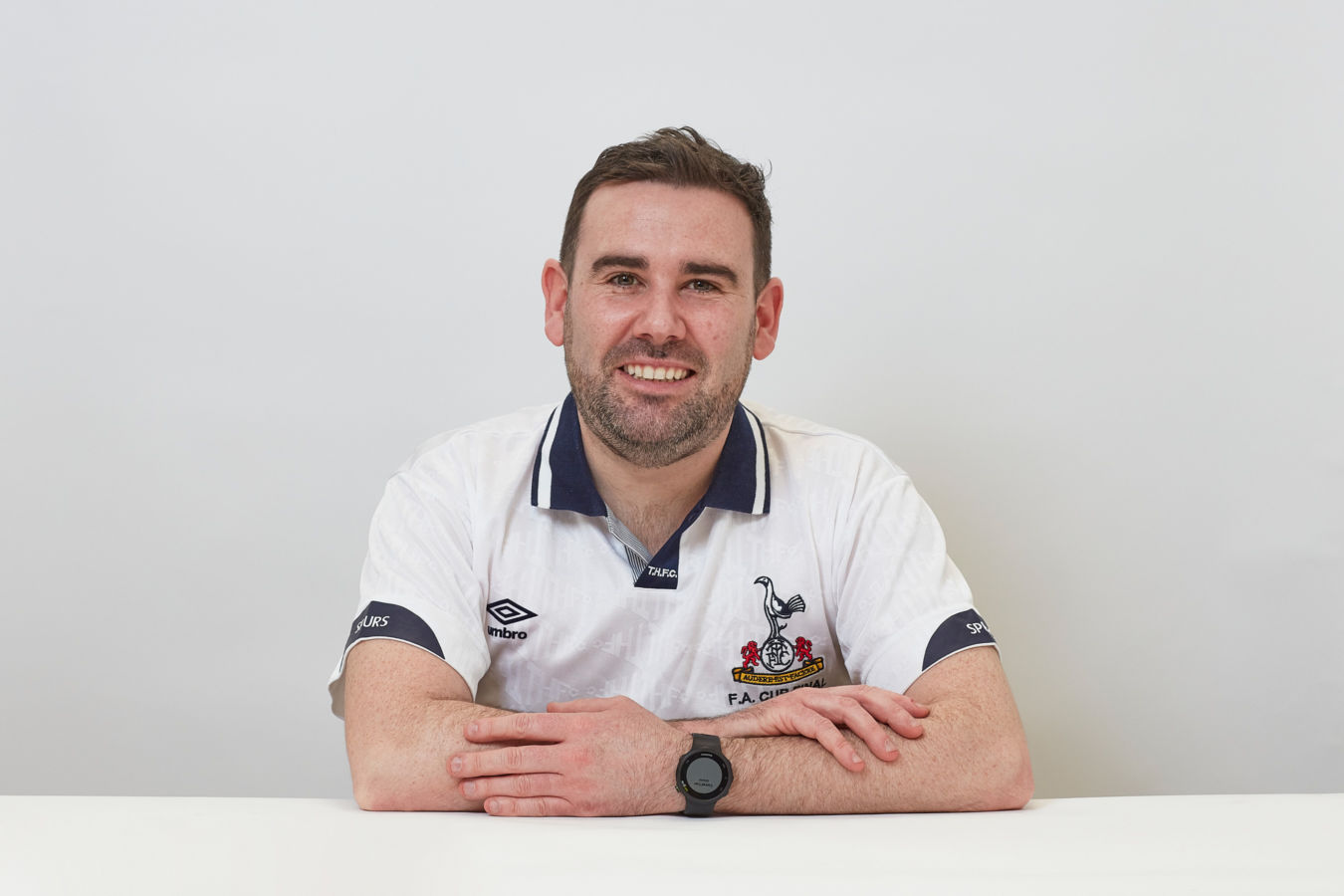 A person with short hair smiles while leaning on a table, wearing a white sports jersey featuring a logo with "F.A. Cup" and "SPURS," and an Umbro emblem, against a plain background.