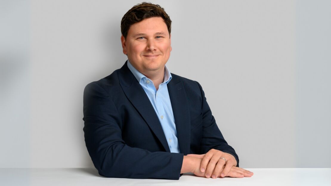 A man in a dark suit and light blue shirt sits at a white table, clasping his hands, against a plain gray background.
