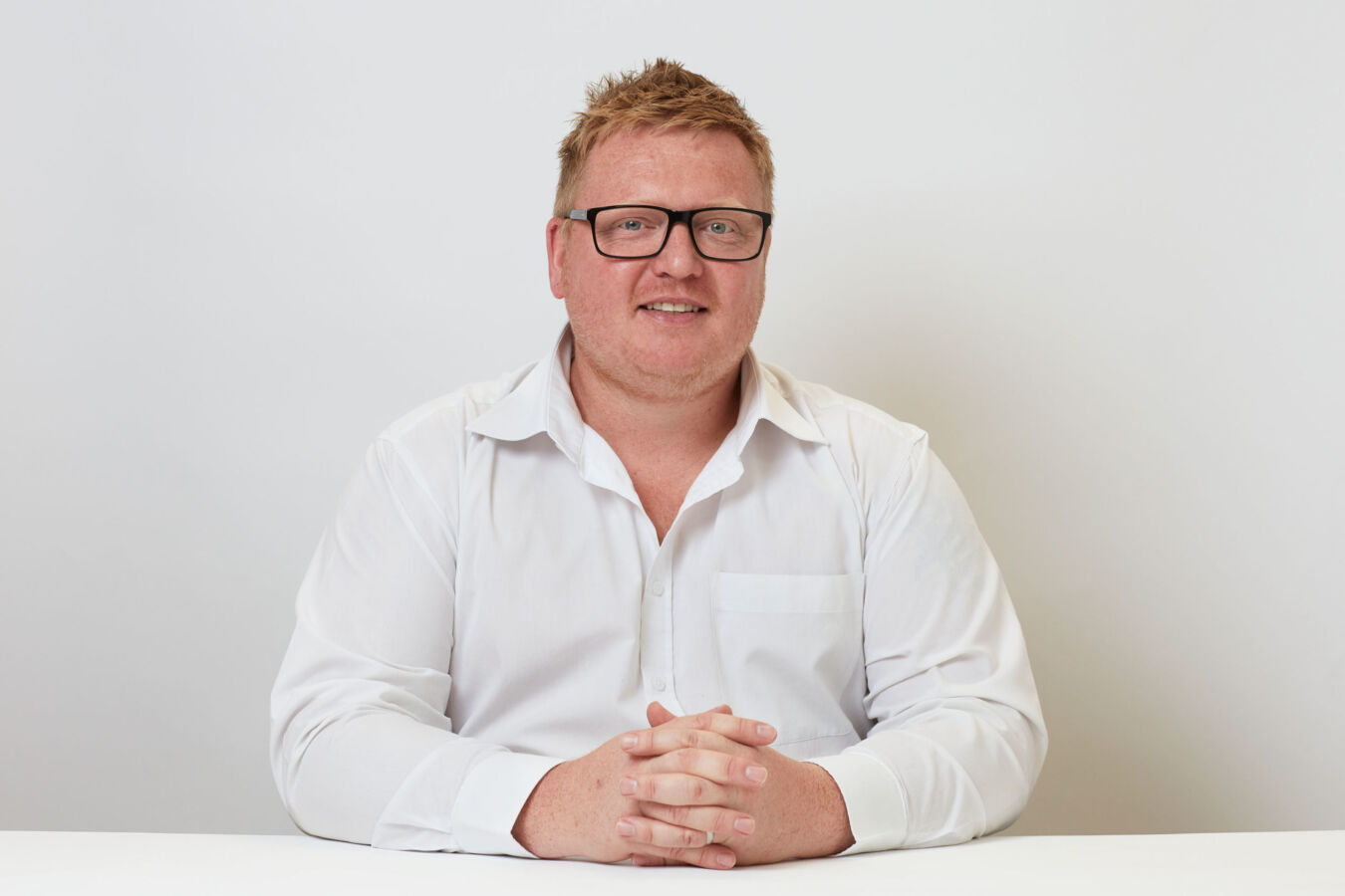 A person in a white shirt sits with clasped hands at a white table against a plain white background.