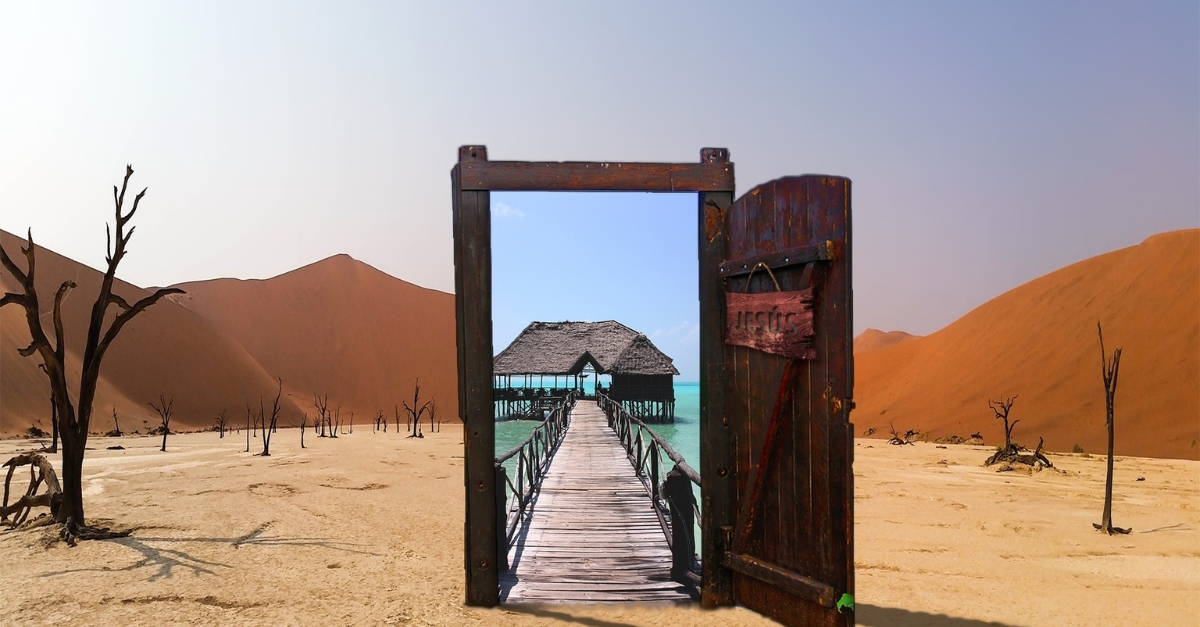 A wooden door stands in a barren desert, opening to a tropical boardwalk over turquoise water. Distant sand dunes and sparse, dead trees contrast with the vibrant seascape beyond.