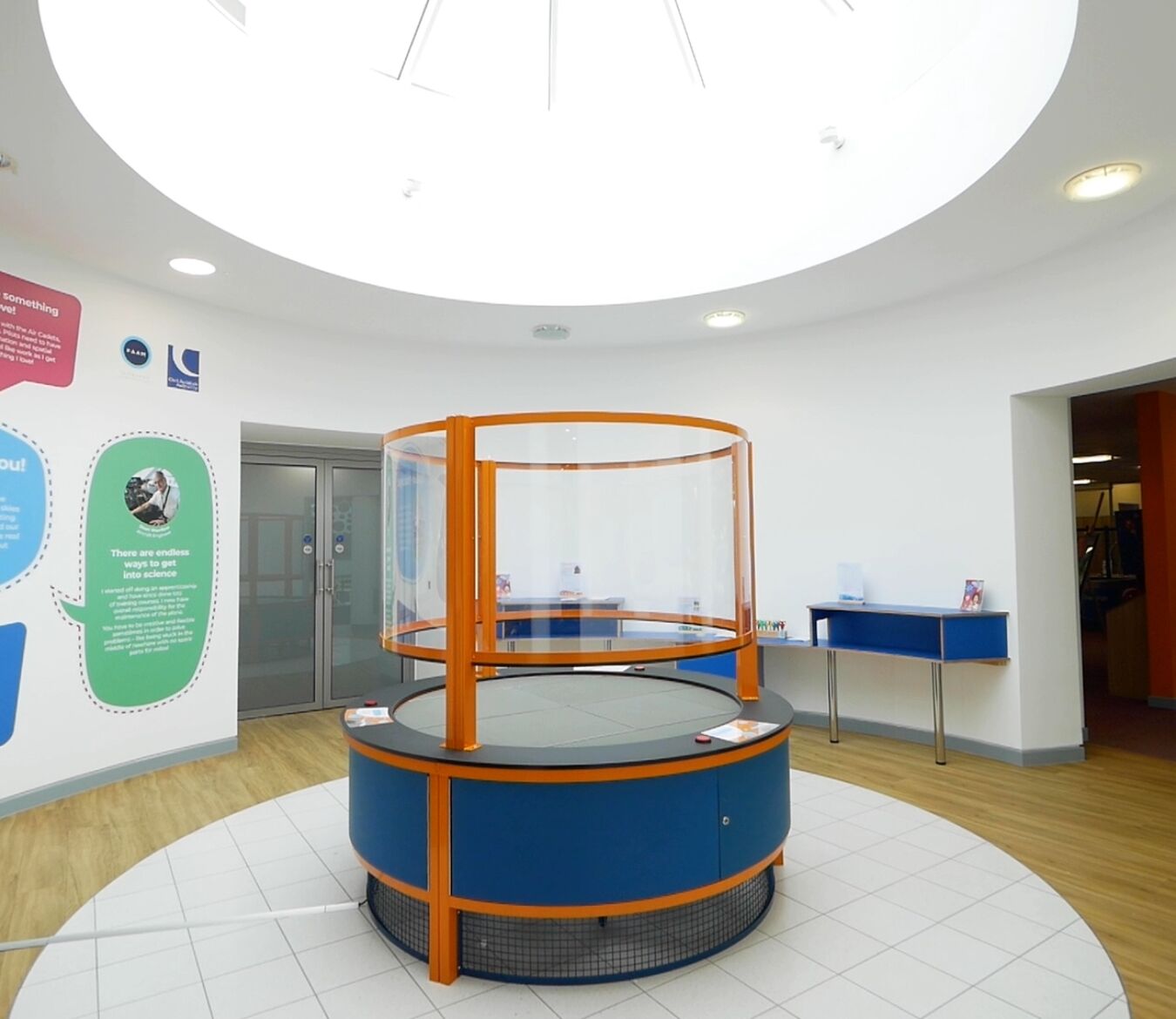 Circular display table with orange frame and clear panels stands unused in a brightly lit lobby. Surrounding walls feature educational text: “Flight Lab – It could be you!” Various educational posters and brochures are visible.