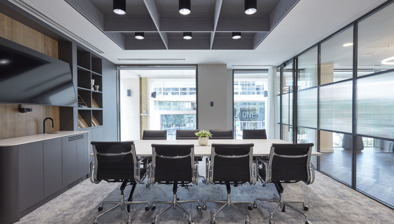 Modern conference room with a long white table, eight black office chairs, floor-to-ceiling windows, and geometric ceiling panels with recessed lighting.