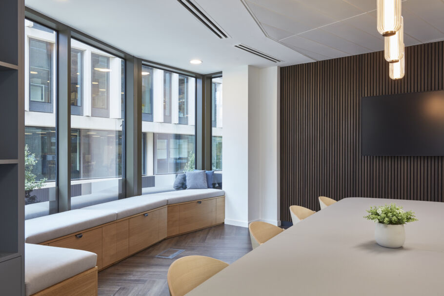 Modern meeting room with large windows, wood paneling, a cushioned window bench, and a long table. Minimalist decor with soft lighting and a plant