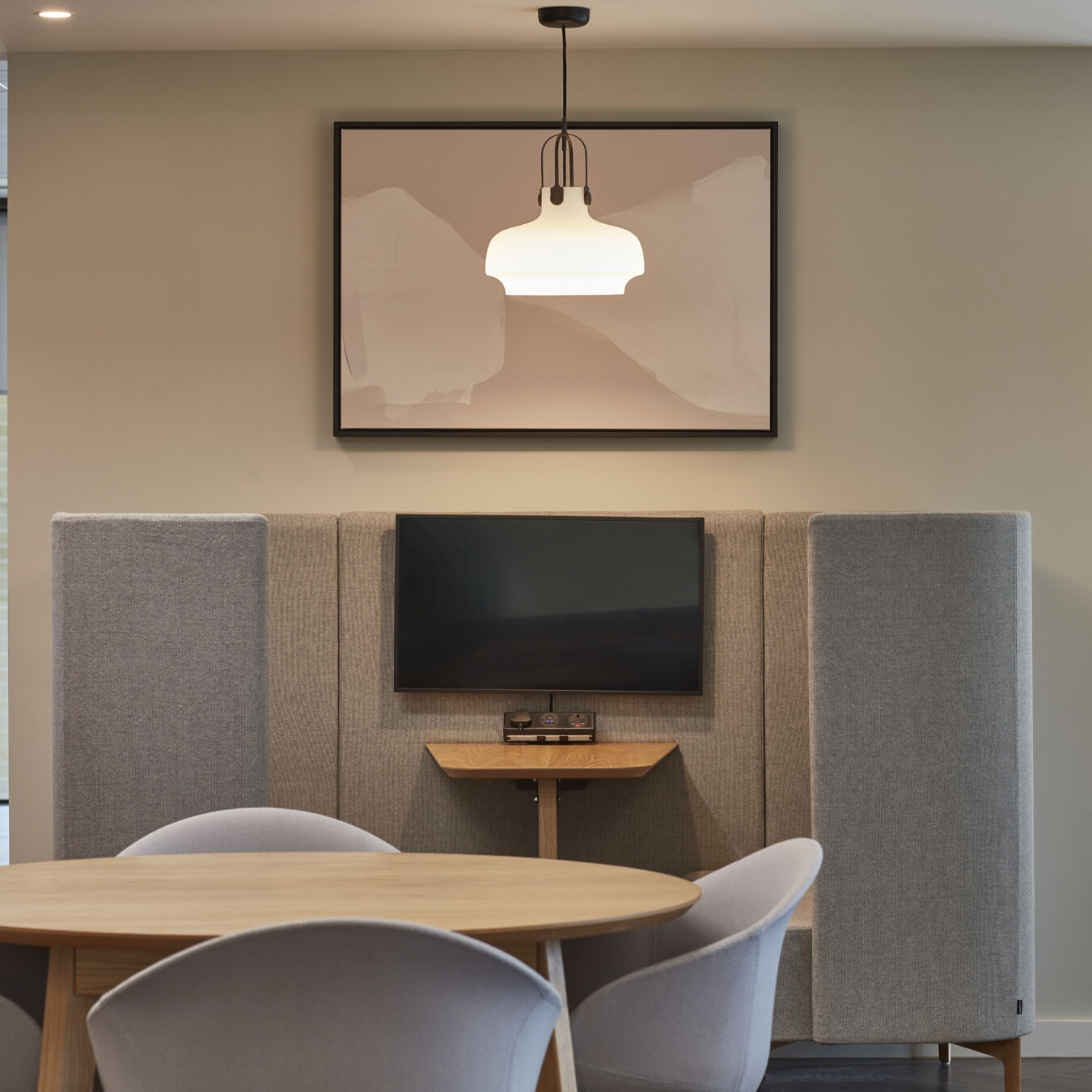 Modern meeting space with a round table and four chairs, a small wall-mounted TV between acoustic panels, and a pendant light hanging above. A framed abstract artwork is displayed on the wall.