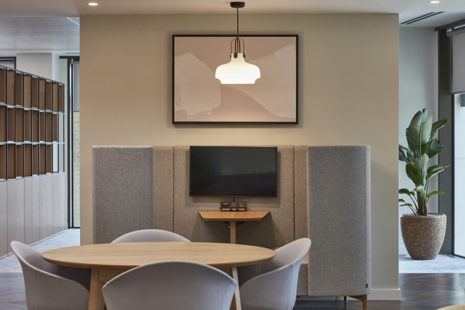 Modern meeting space with a round table and four chairs, a small wall-mounted TV between acoustic panels, and a pendant light hanging above. A framed abstract artwork is displayed on the wall.