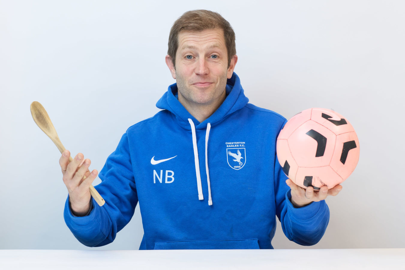A person in a blue hoodie, labeled "Chesterton Eagles F.C." and "NB," holds a wooden spoon in one hand and a pink soccer ball in the other, against a plain background.