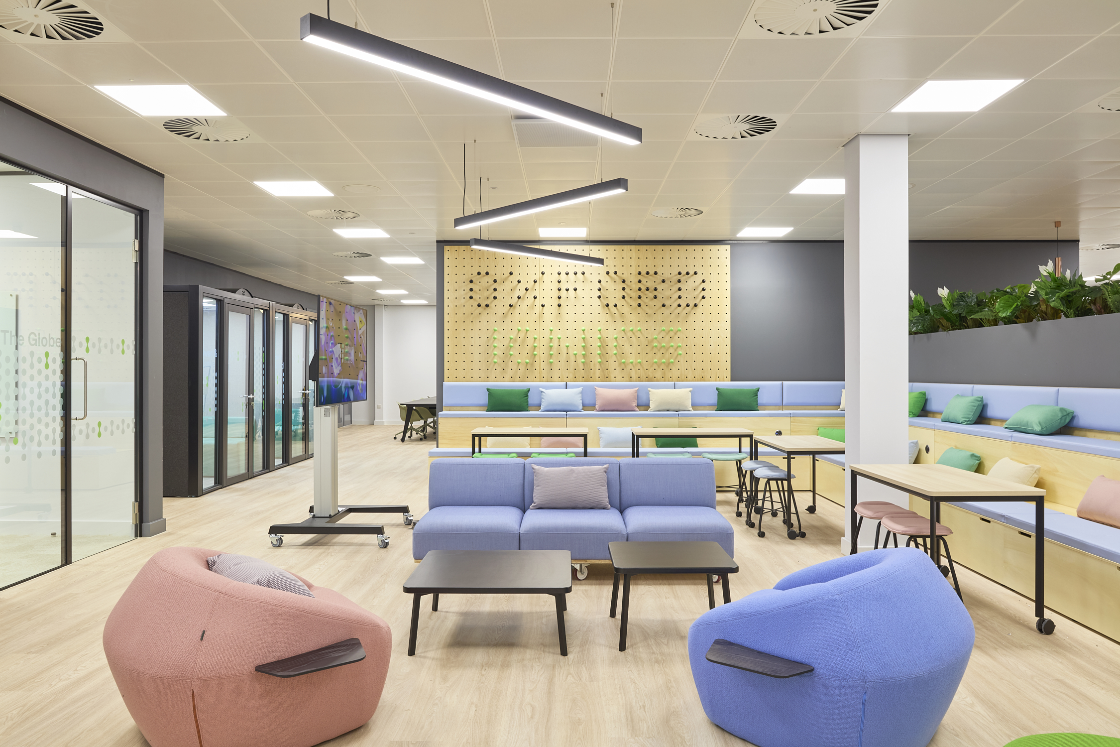 Modern office lounge with pastel furniture, including blue sofas and pink beanbag chairs, arranged around tables. Pegboard wall with plants and text "The Globe" visible through glass door.