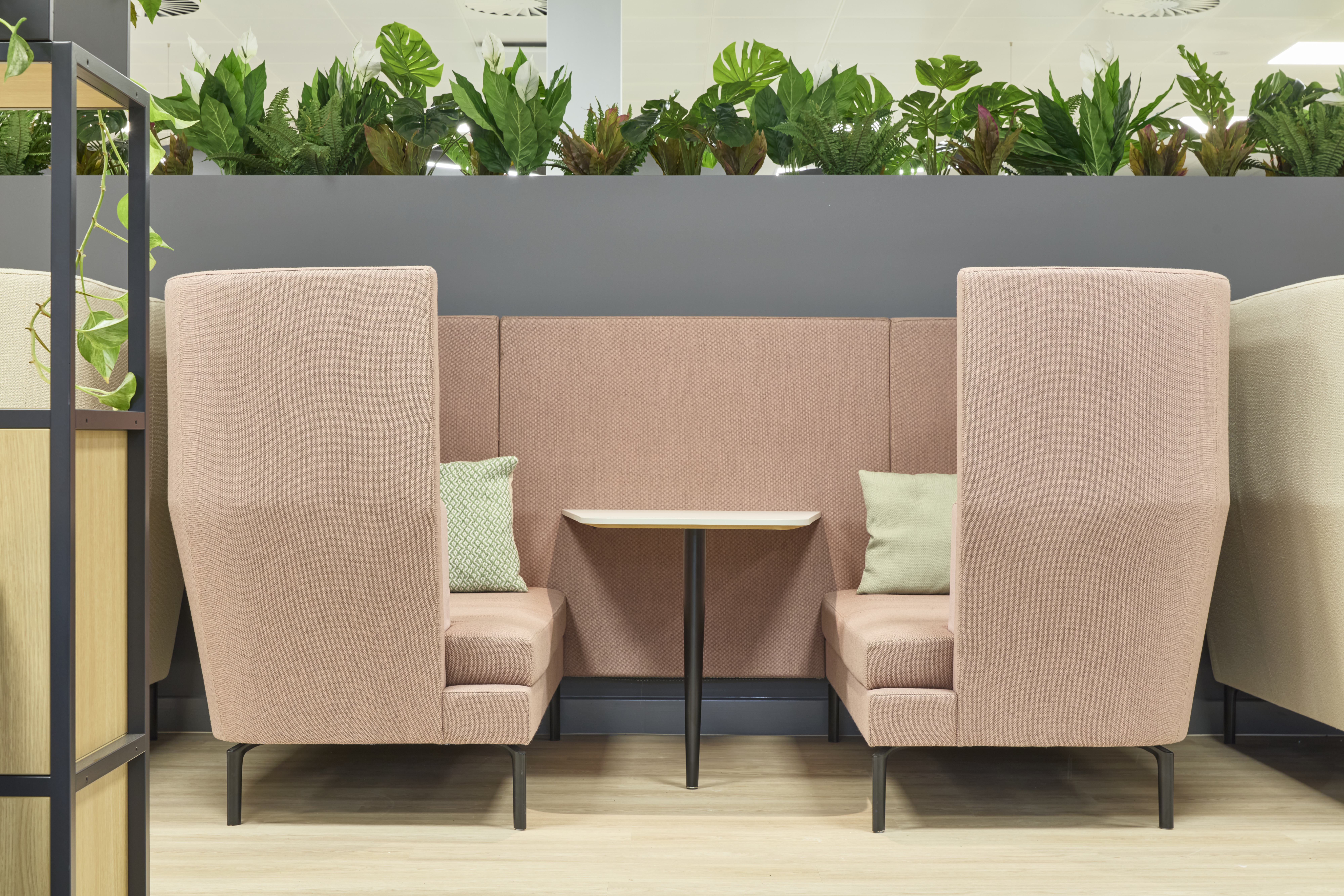 Pink upholstered booth seating with a central table is set against a background of green plants on a shelf, creating a cozy, modern interior space.
