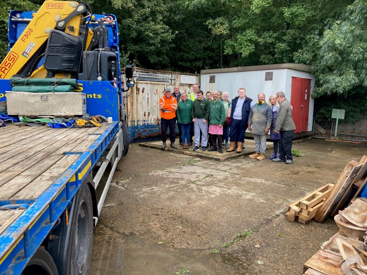 A group of people stand together beside a large truck with a crane in a wooded, industrial area, near a rusty container and a small building with a red door.