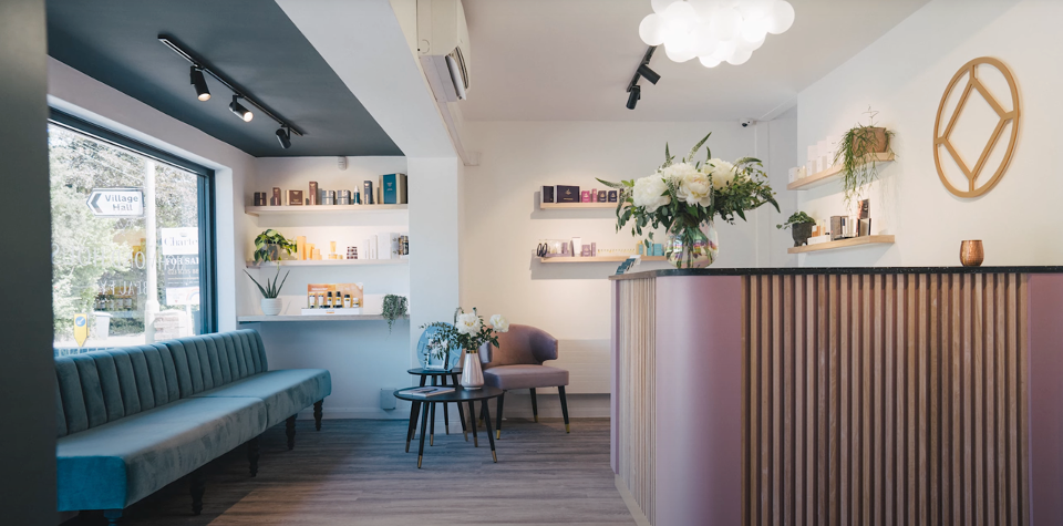 Reception area features a sleek counter with floral arrangement; shelves display beauty products against white walls. A modern sofa rests by a large window, with a circular decorative wall piece.