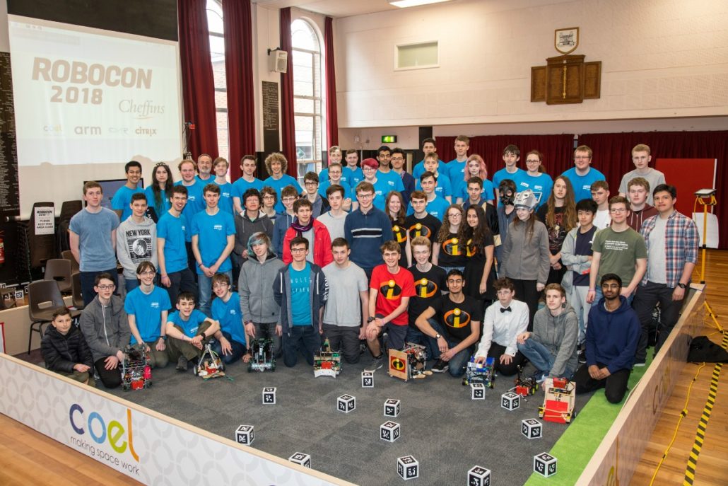 A large group of people poses with robotics projects on a stage, surrounded by audience seating. A banner reads "ROBOCON 2018" with sponsor logos. The atmosphere is enthusiastic and collaborative.