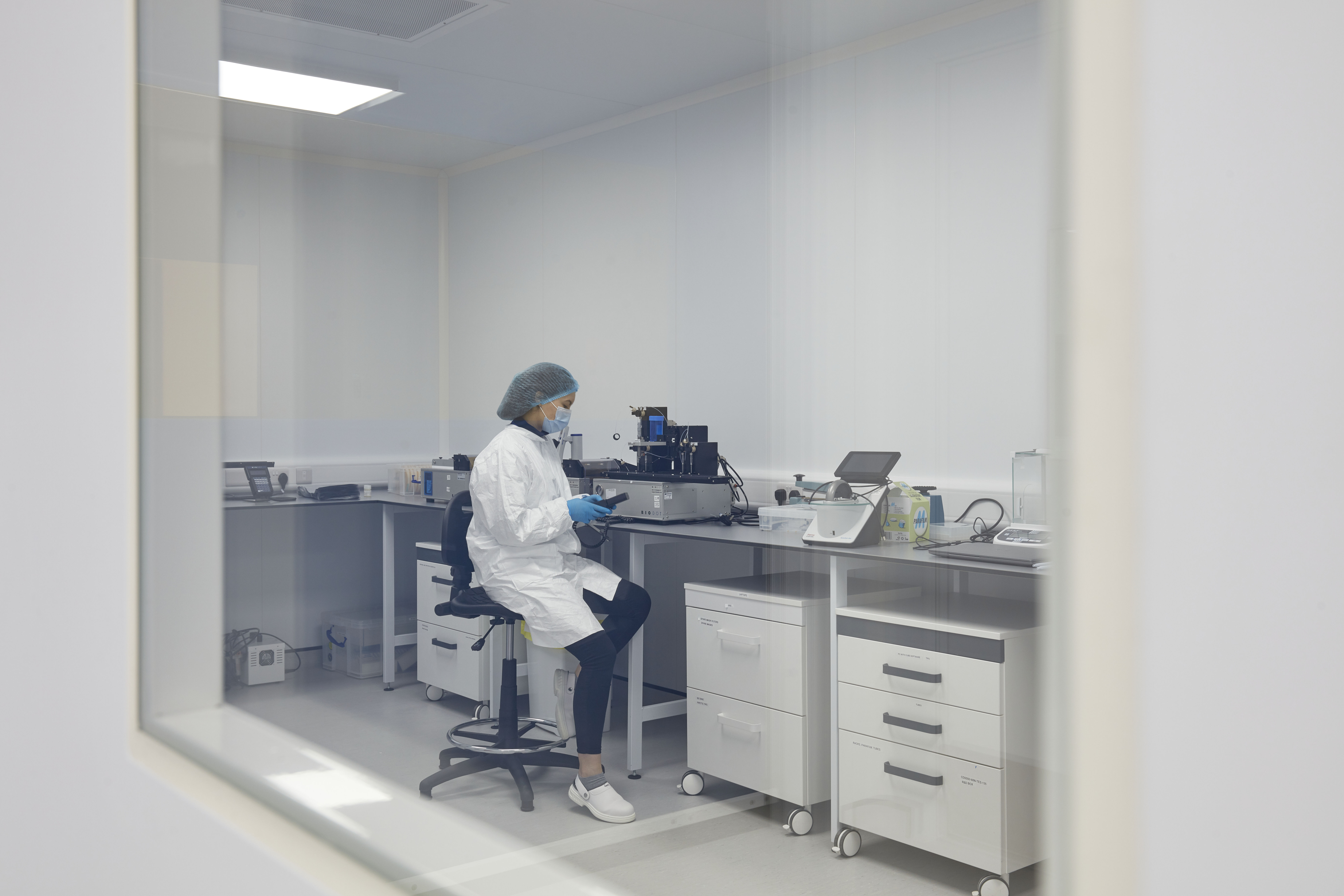 A lab technician, wearing protective attire and gloves, works with a device on a counter in a sterile lab environment with white cabinets and equipment.