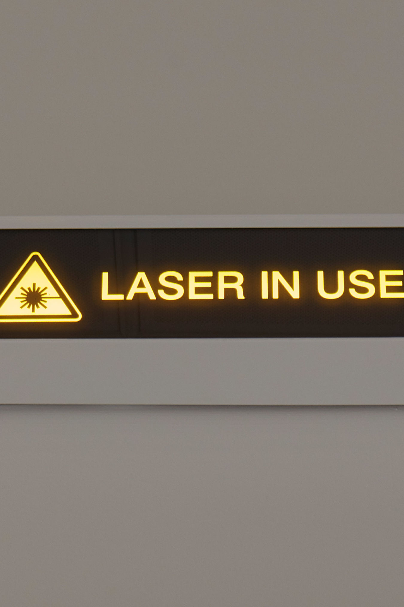 Illuminated sign displaying the text "LASER IN USE" with a laser warning symbol, set against a neutral wall background.