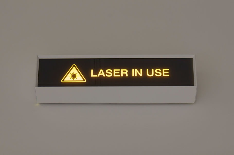 Illuminated sign displaying the text "LASER IN USE" with a laser warning symbol, set against a neutral wall background.
