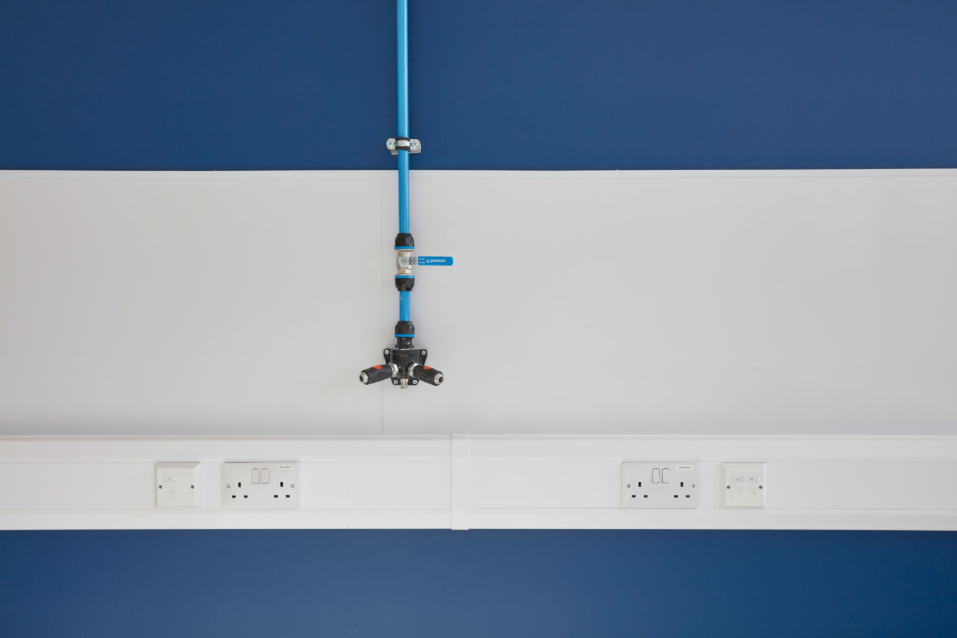A blue pipe system with multiple fittings is mounted on a white and blue wall, featuring the text "prevost" and outlets below.