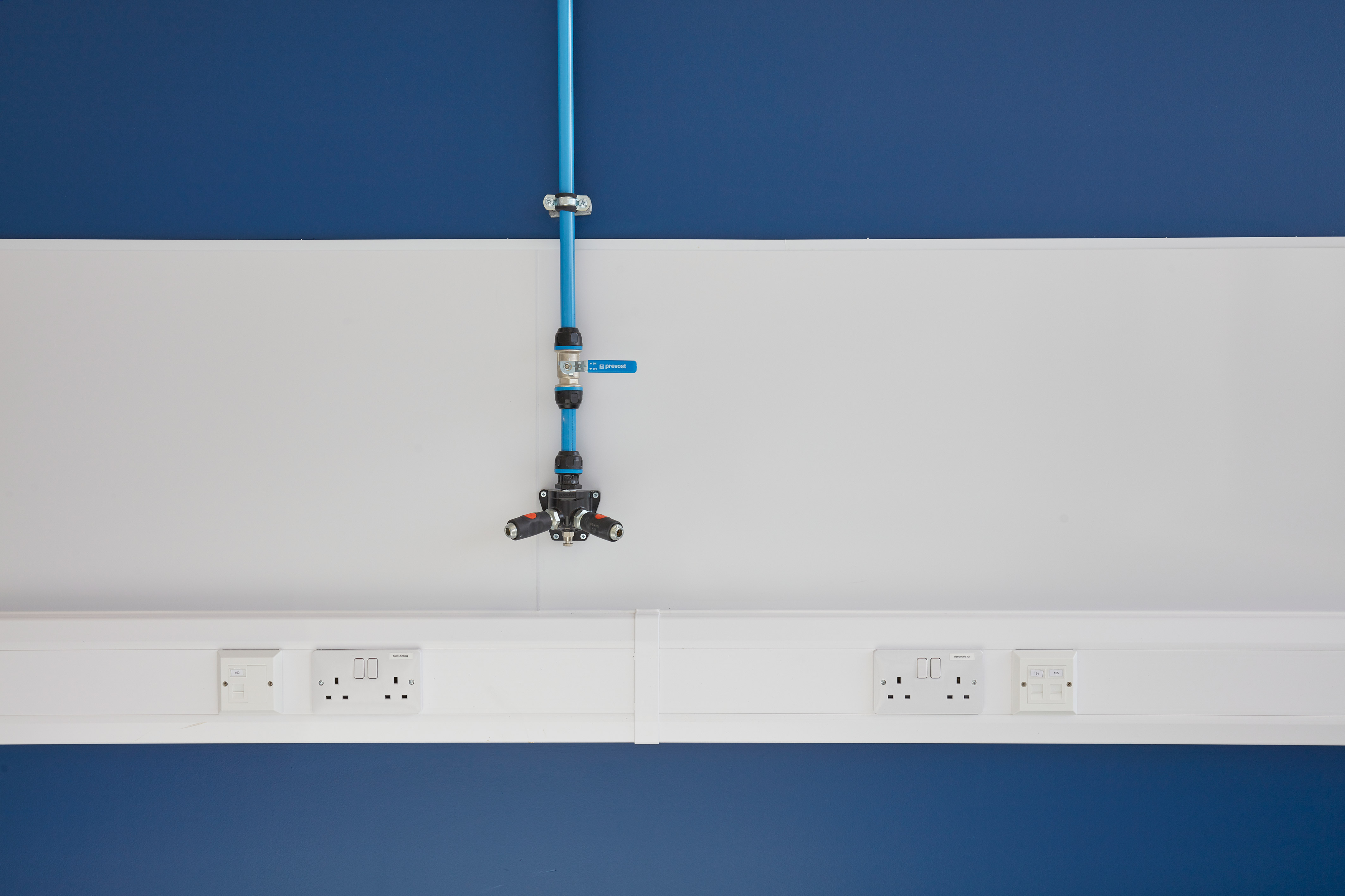 A blue pipe system with multiple fittings is mounted on a white and blue wall, featuring the text "prevost" and outlets below.