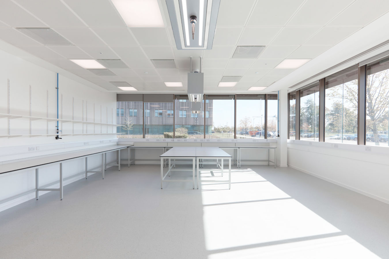Laboratory room with empty white workbenches and tables, illuminated by natural light from large windows with an outdoor view of a building and trees in a professional setting.