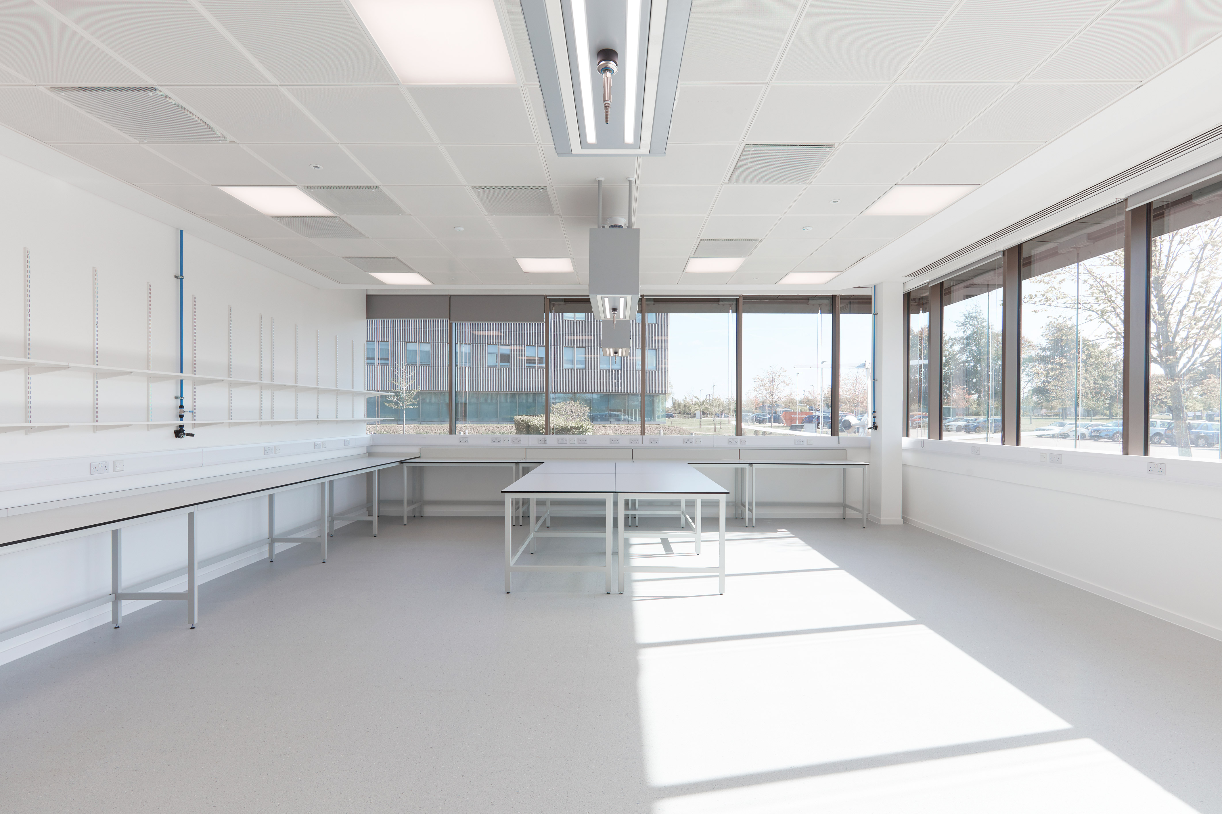 Laboratory room with empty white workbenches and tables, illuminated by natural light from large windows with an outdoor view of a building and trees in a professional setting.
