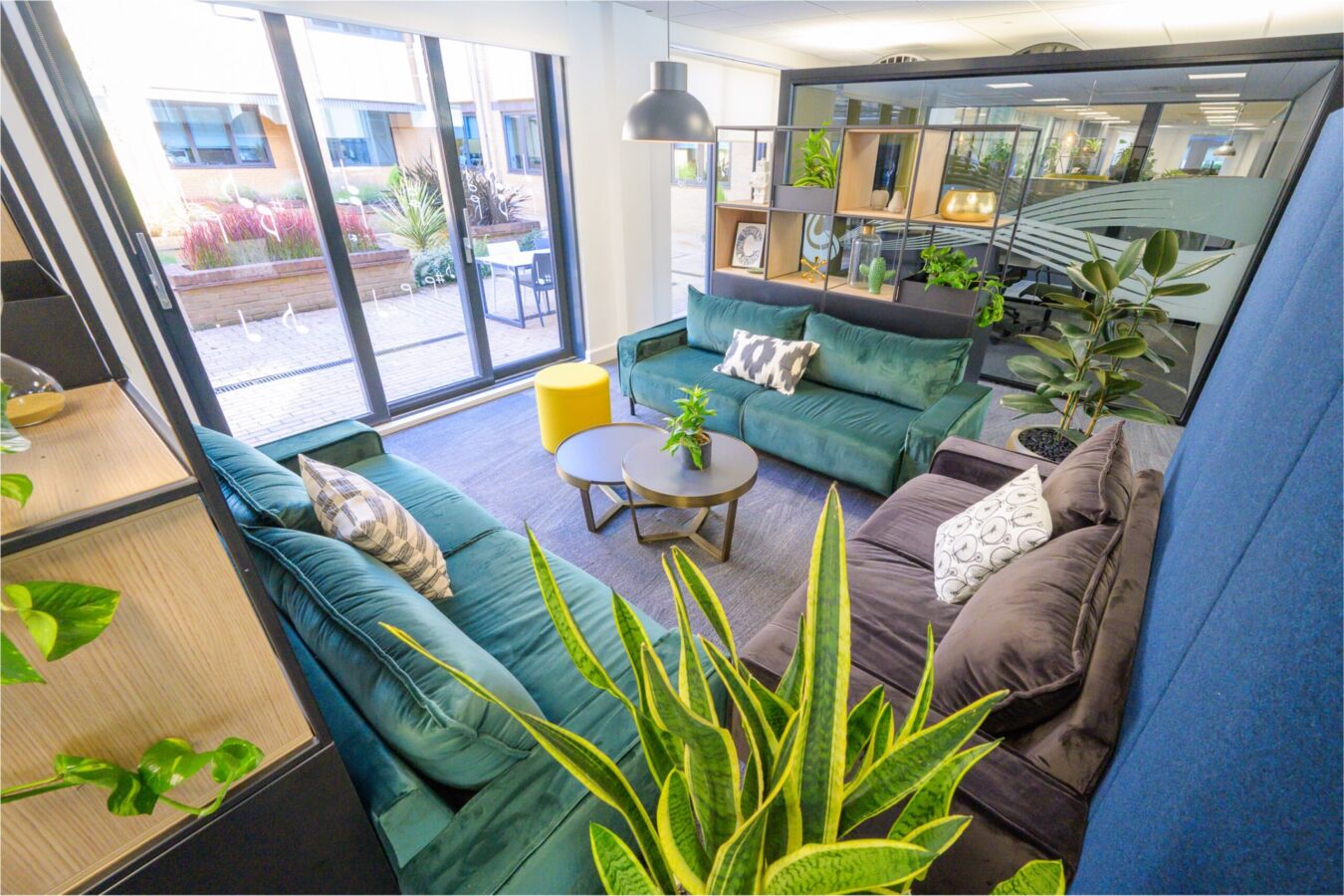 Modern lounge area with green and teal sofas, indoor plants, and large glass doors opening to a patio.