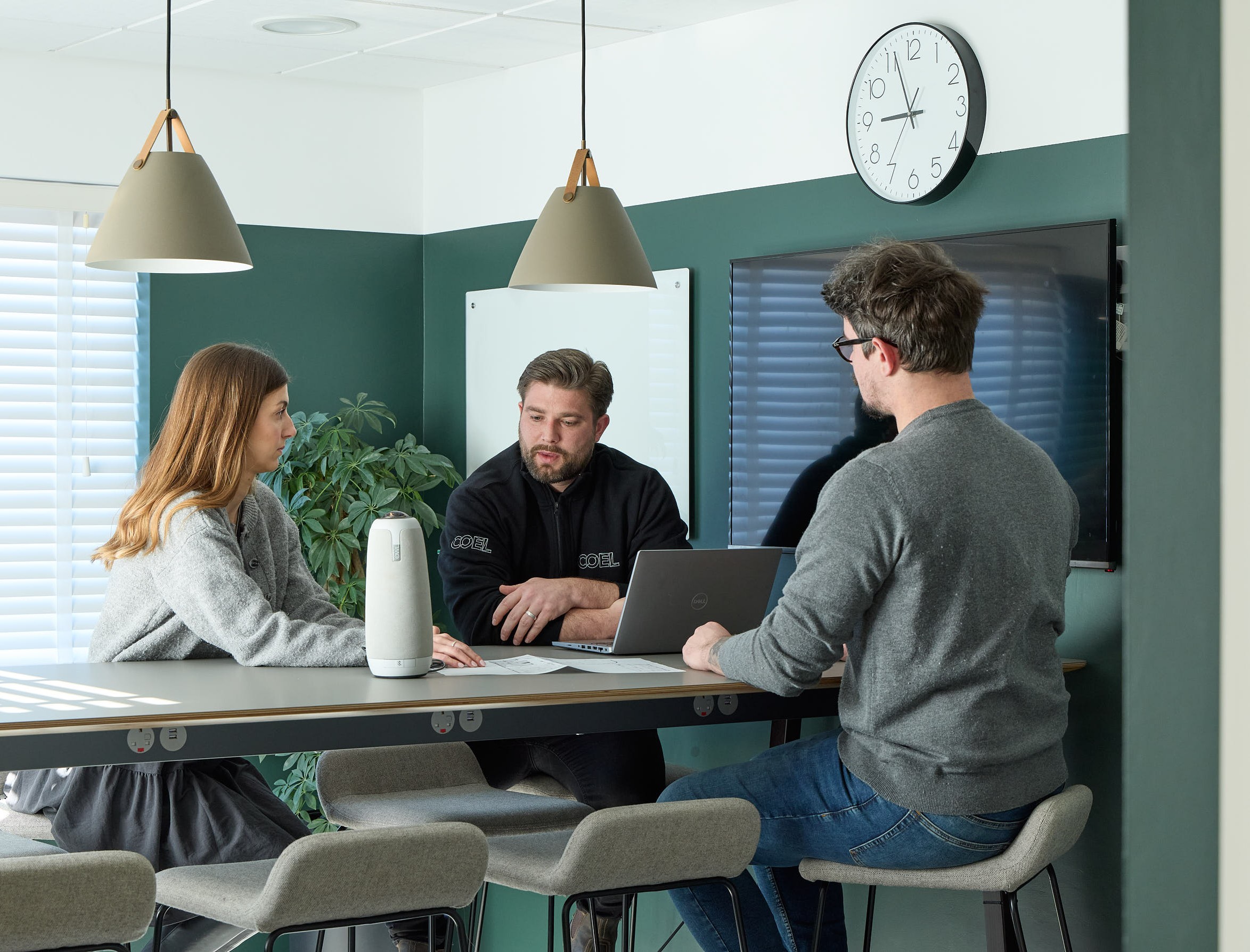 Three people converse around a desk in a modern office, with laptops and notes. The atmosphere is collaborative, under soft pendant lights and a wall clock