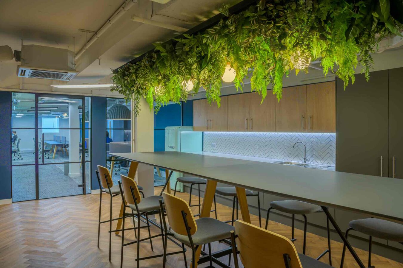 A modern office kitchen with a long table and high chairs beneath a ceiling adorned with hanging plants and lights, adjacent to wooden cabinets and an open workspace visible through glass doors.