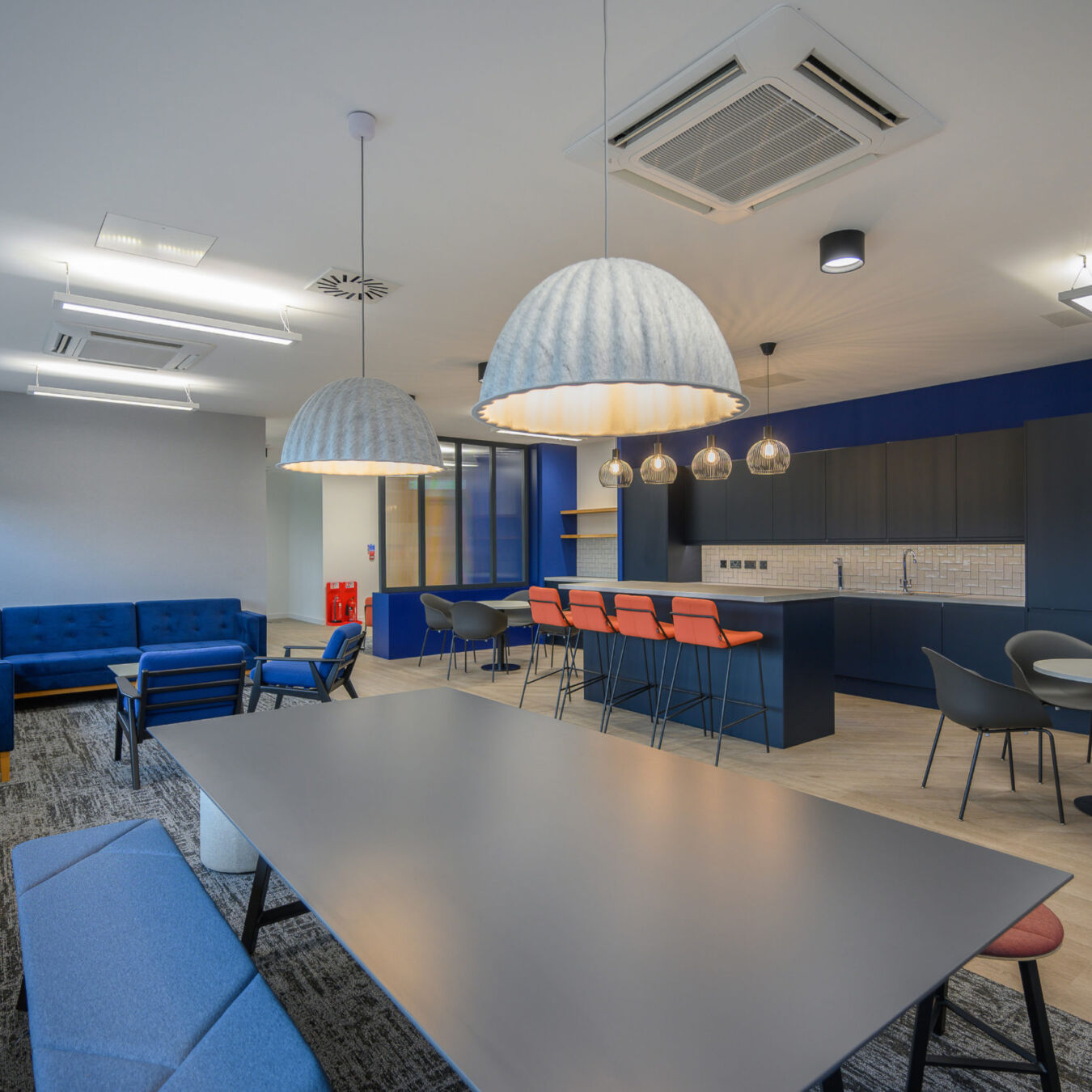 Modern office kitchen and breakout area featuring large pendant lights, communal tables, blue and red seating, and a kitchenette with overhead cabinets