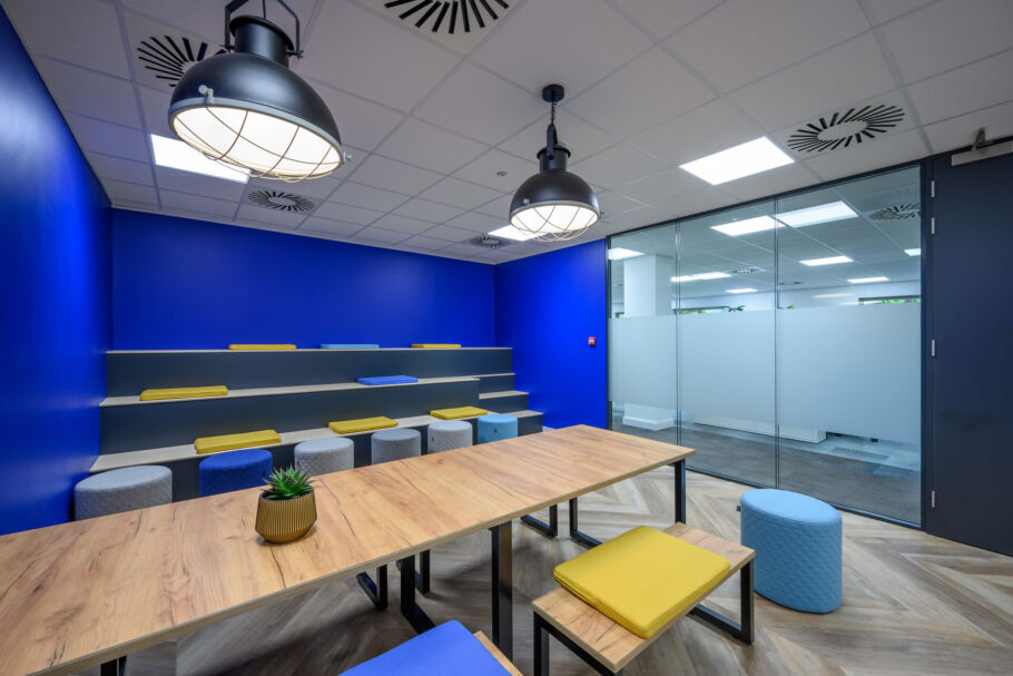 Modern meeting room with blue walls and tiered seating featuring yellow and blue cushions. Wooden table with small potted plant; bright, open atmosphere.