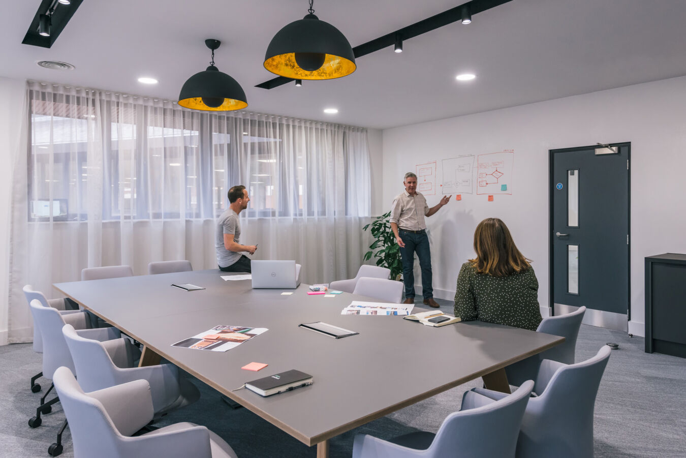 Modern meeting room with a large table, pendant lighting, and three people engaged in a collaborative discussion while one presents at a whiteboard.