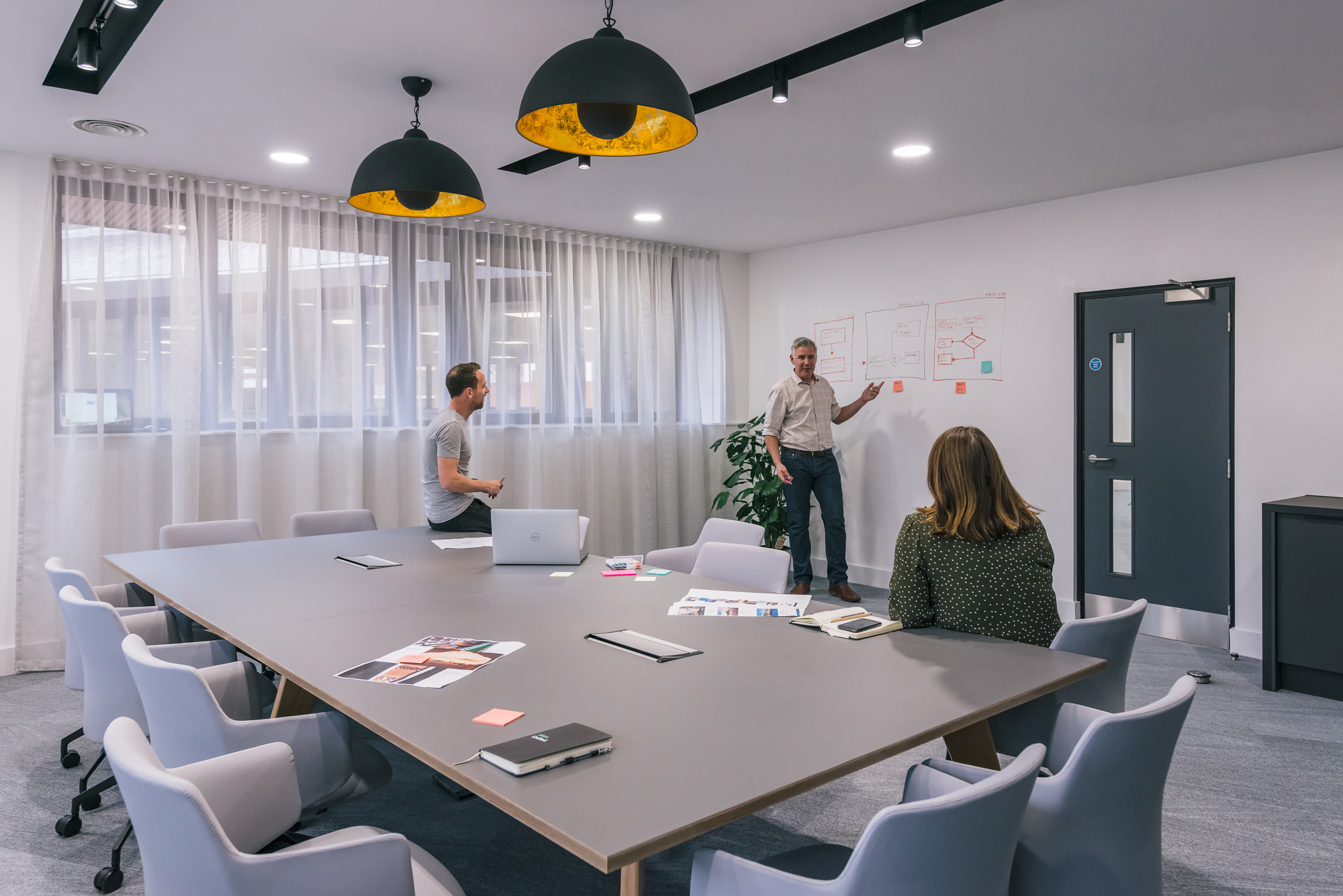 Modern meeting room with a large table, pendant lighting, and three people engaged in a collaborative discussion while one presents at a whiteboard.
