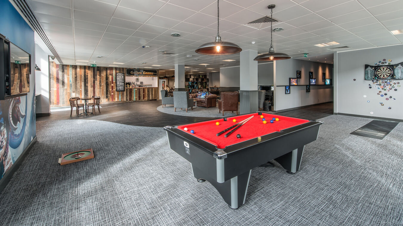 A red pool table with balls and cues is placed in a spacious recreational room featuring a dartboard, lounge chairs, a bar labeled "Golden Gnome," and artwork on the walls.