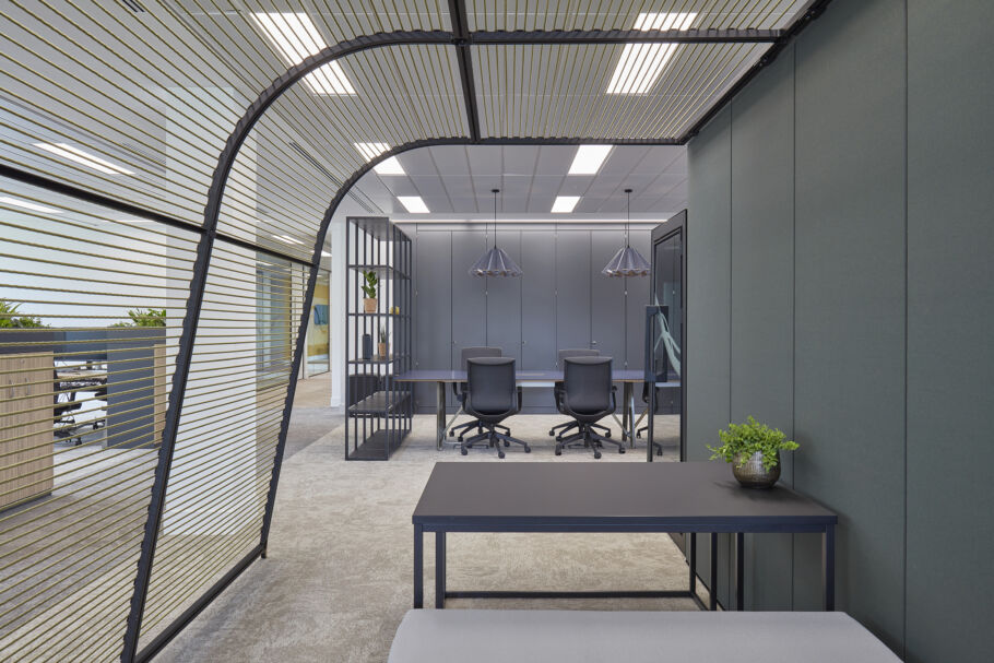 Modern office space featuring tables with chairs facing a wall with cabinets. Overhead lights illuminate the area. A plant decorates a table in the foreground next to a metal partition wall.