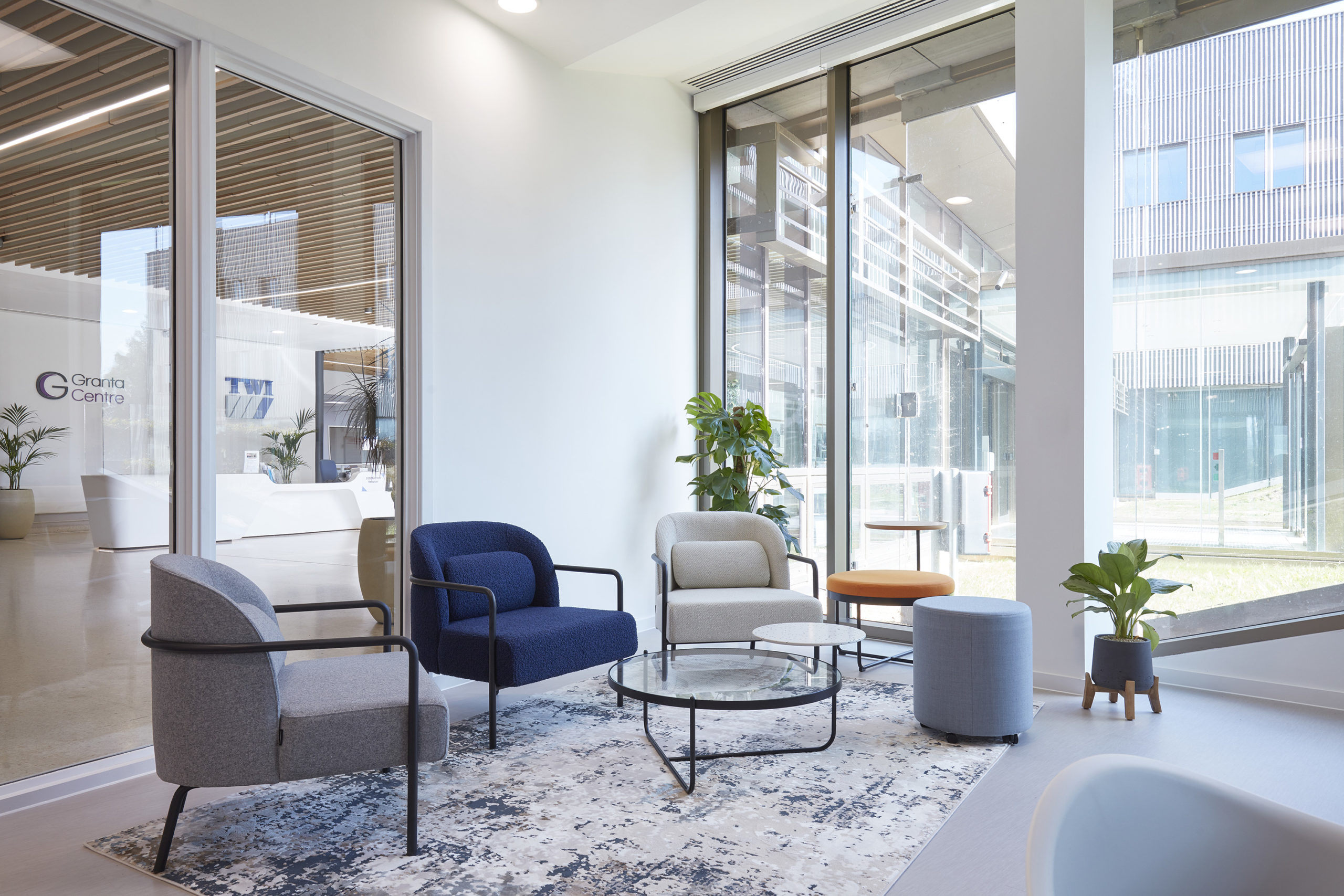 Seating area with modern chairs surrounds a glass coffee table, situated by large windows in a bright lobby. Text on door reads "Granta Centre." Potted plants add greenery.
