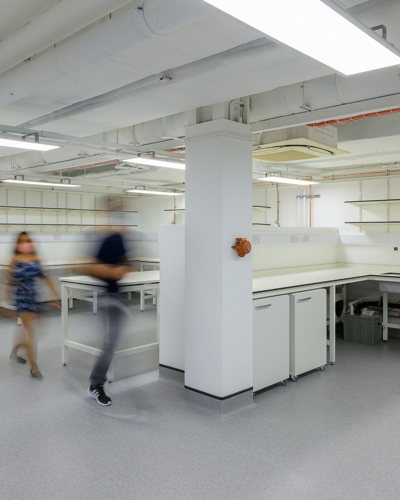 Spacious laboratory with white workbenches, overhead shelving, and exposed ceiling pipes, with two people walking through the area creating motion blur.