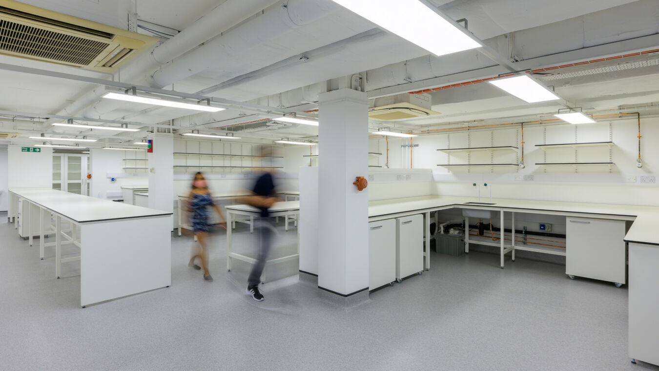 Spacious laboratory with white workbenches, overhead shelving, and exposed ceiling pipes, with two people walking through the area creating motion blur.