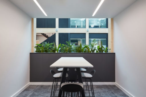 A modern office space with a white table and four black chairs. Behind the table, there are green plants in front of a large window, creating a fresh and inviting atmosphere.