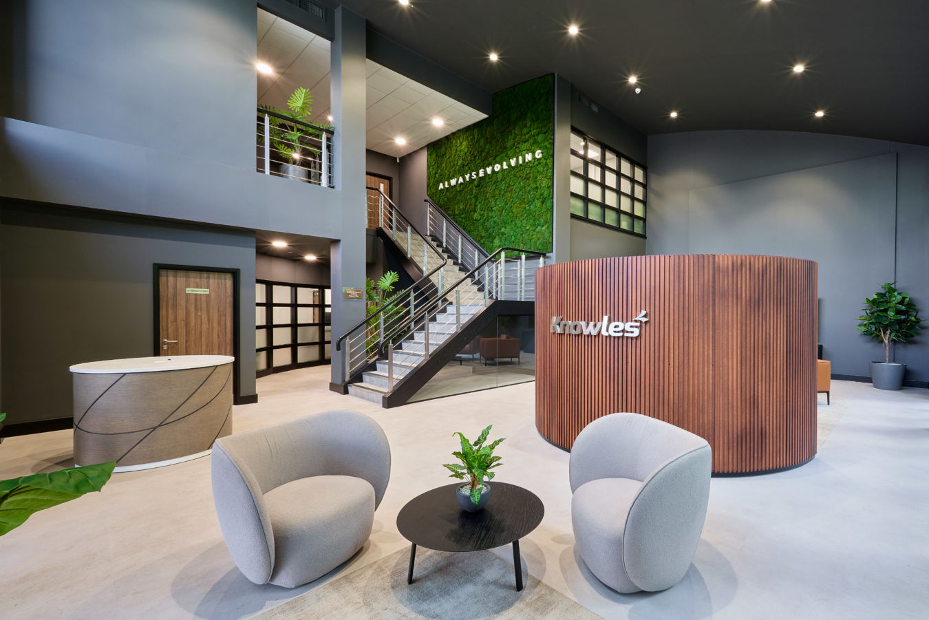 Modern office lobby with two curved chairs, a round table with a plant, and a wooden reception desk. Stairs lead to a green