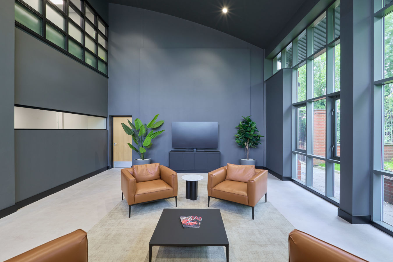 Modern lounge with high ceilings, featuring tan leather chairs, a black coffee table, large windows, and potted plants. The atmosphere is sleek and inviting.