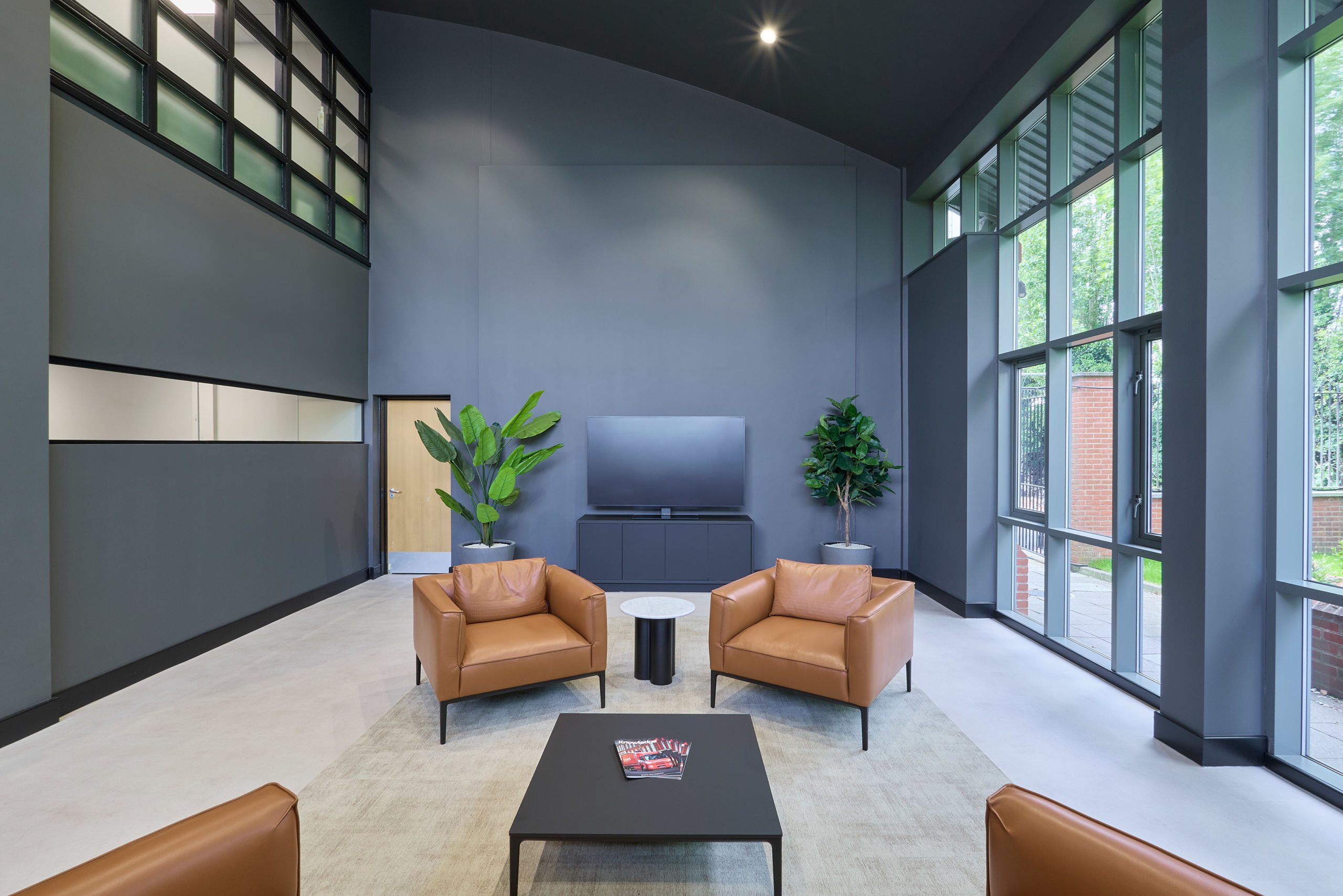 Modern lounge with high ceilings, featuring tan leather chairs, a black coffee table, large windows, and potted plants. The atmosphere is sleek and inviting.
