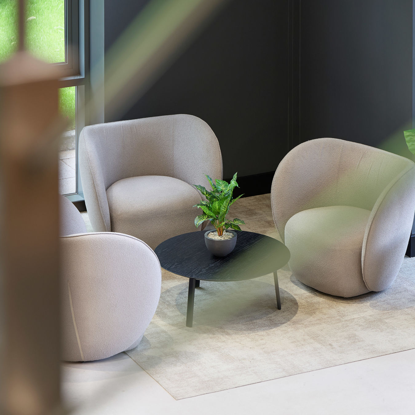 Cozy seating area with three beige armchairs around a black round table holding a potted plant. Soft lighting and a neutral-toned ambiance.