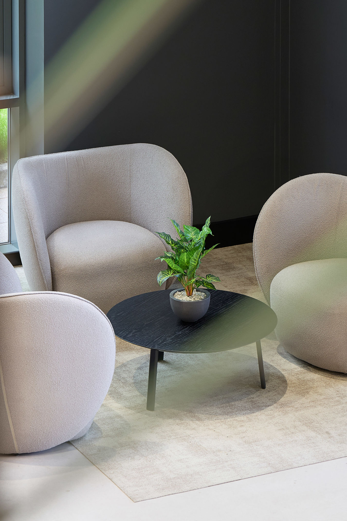 Cozy seating area with three beige armchairs around a black round table holding a potted plant. Soft lighting and a neutral-toned ambiance.