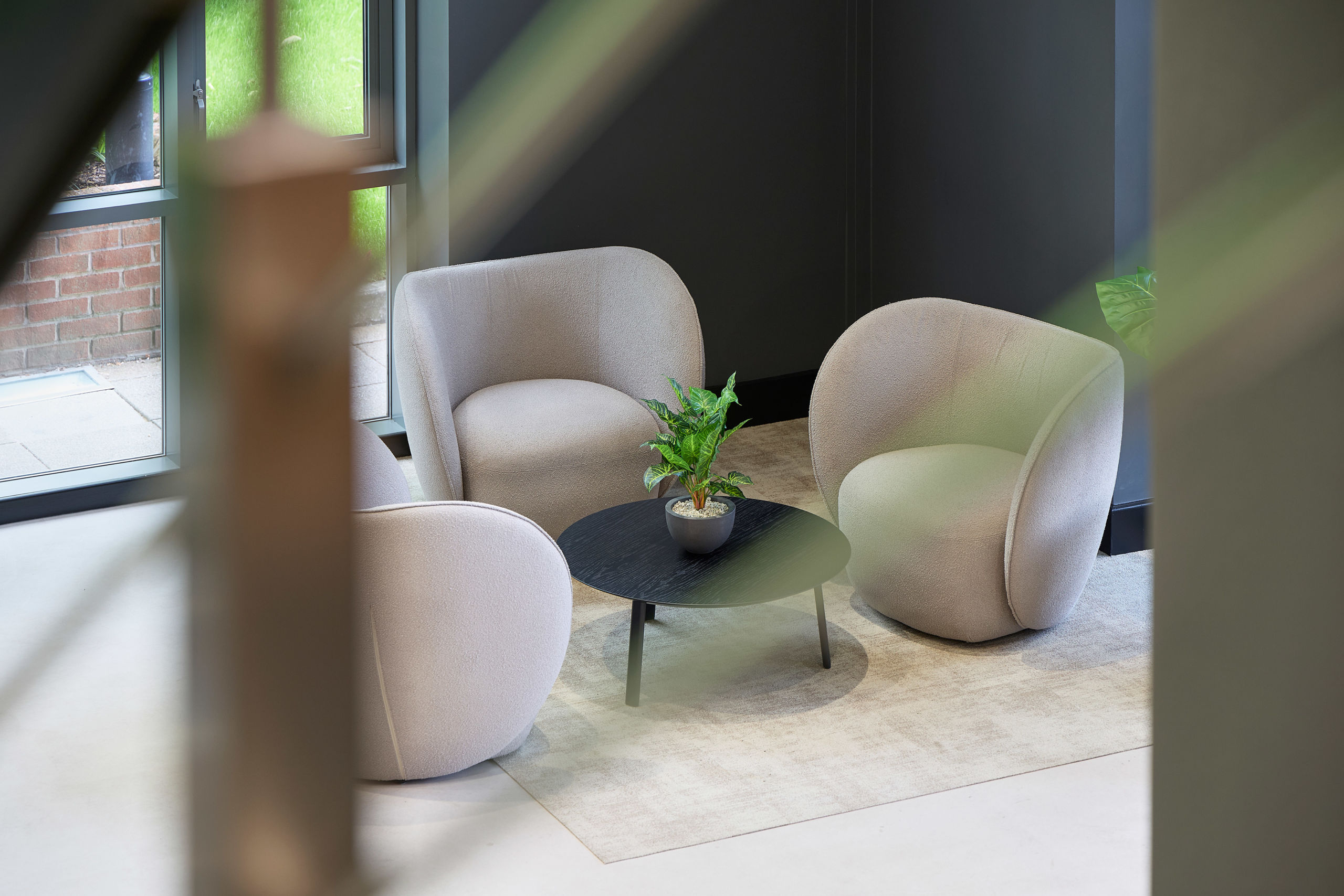 Cozy seating area with three beige armchairs around a black round table holding a potted plant. Soft lighting and a neutral-toned ambiance.