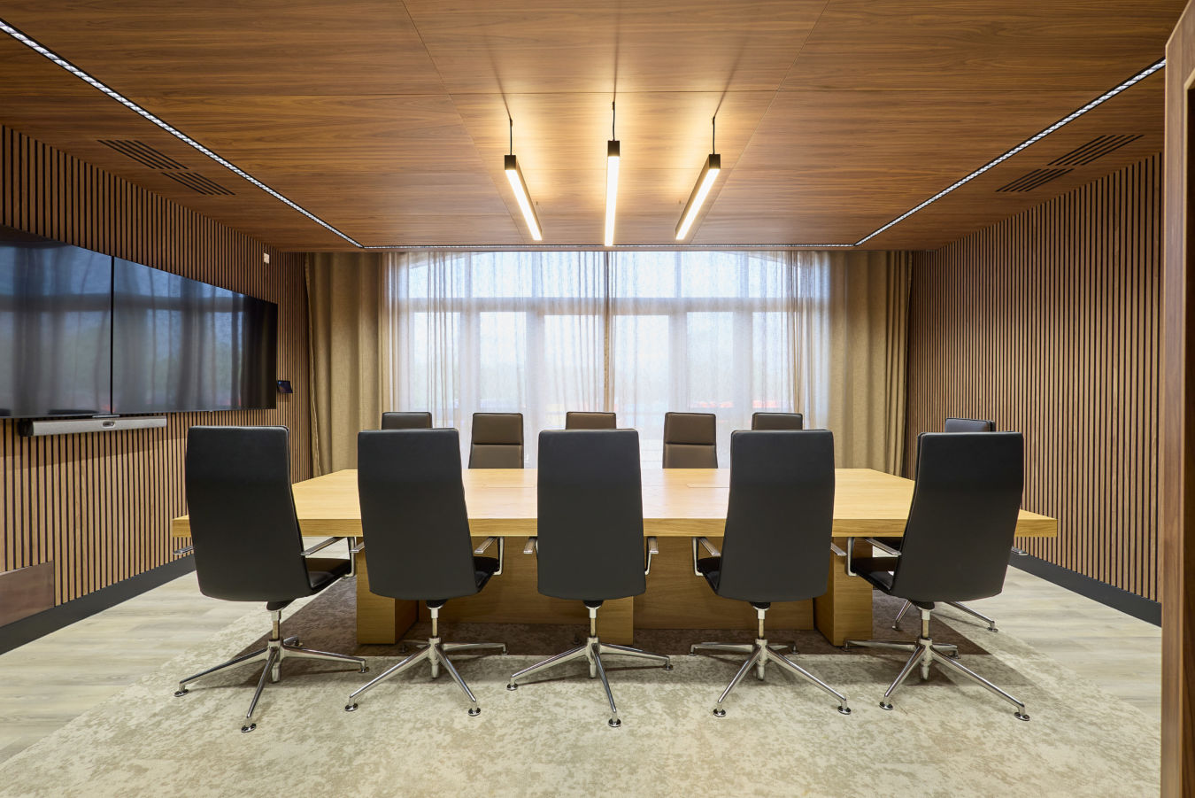 Modern conference room with a large wooden table surrounded by eight black chairs. Warm lighting, wooden walls, and a flat-screen TV create a professional atmosphere.
