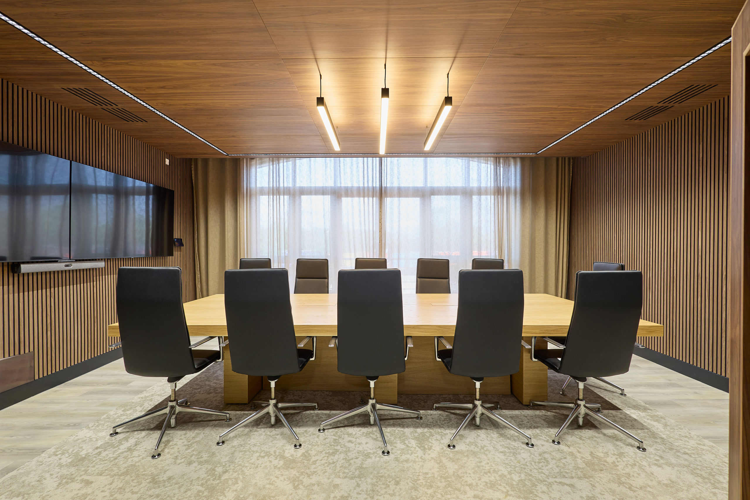 Modern conference room with a large wooden table surrounded by eight black chairs. Warm lighting, wooden walls, and a flat-screen TV create a professional atmosphere.