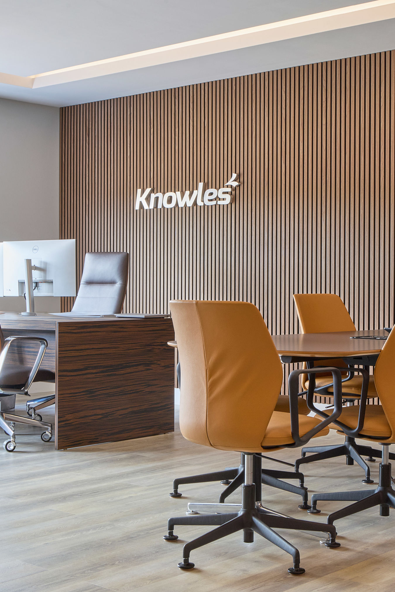 Modern office space with brown leather chairs around a conference table, large TV on a wood-paneled wall, and large windows allowing natural light.