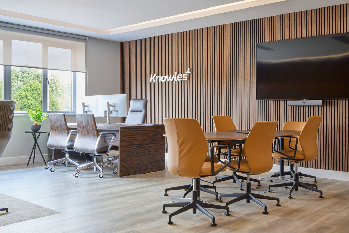 Modern office space with brown leather chairs around a conference table, large TV on a wood-paneled wall, and large windows allowing natural light.